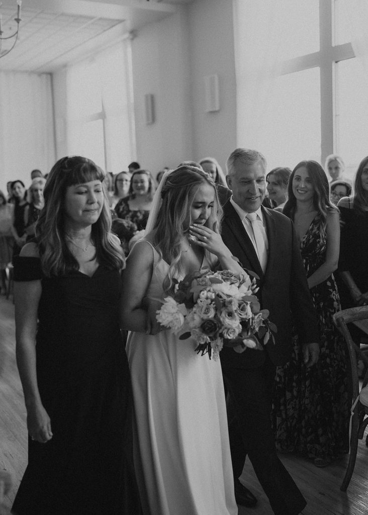 black and white image of bride crying walking down the aisle with her parents