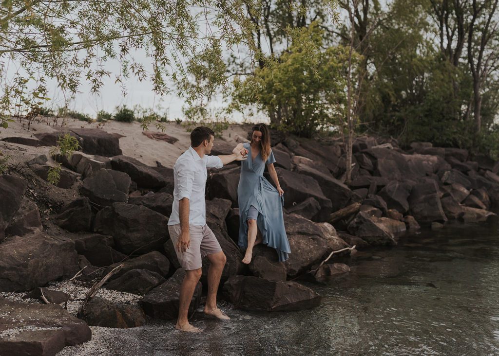 Fiance helping his girlfriend down off some rocks at the beach in rochester ny during their engagement session