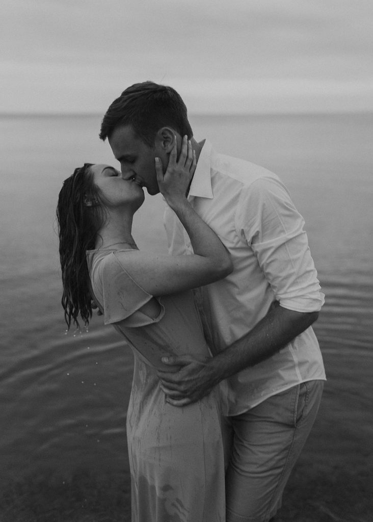 black and white photo of engaged couple standing in lake erie kissing during their engagement session