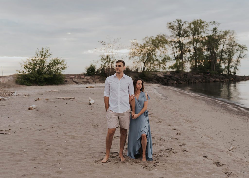 couple standing side by side while holding hands and looking in different direction down the beach