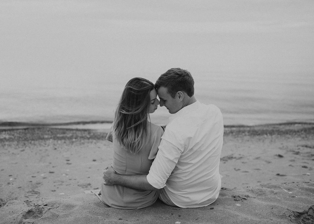 black and white photo of couple sitting on the beach and the photographer in back of them