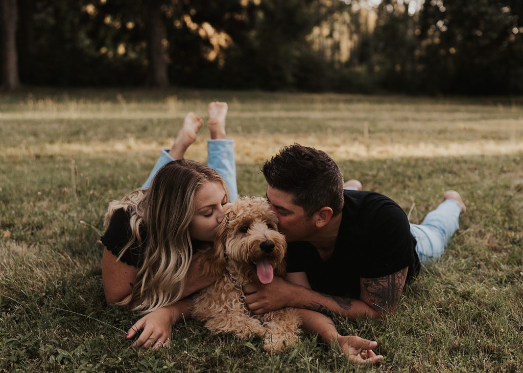 pet parents kissing their golden doodle as they all lay on the grass with their furry friend in the middle