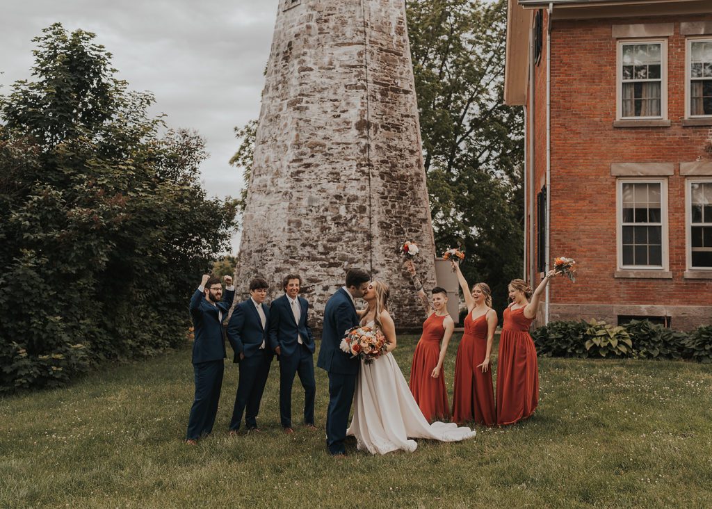bride and groom kissing outside of the charolette lighthouse in rochester ny with bridal party cheering