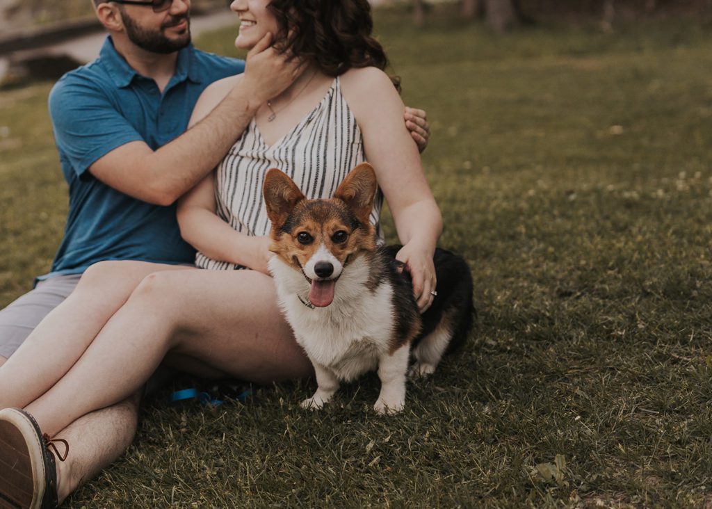 photo of couple smiling in the background but the main focus of the camera is on their pet corgi who is looking at the camera with his tongue out