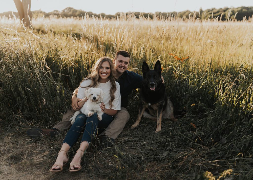 engaged couple sitting in a field with their two dogs. 