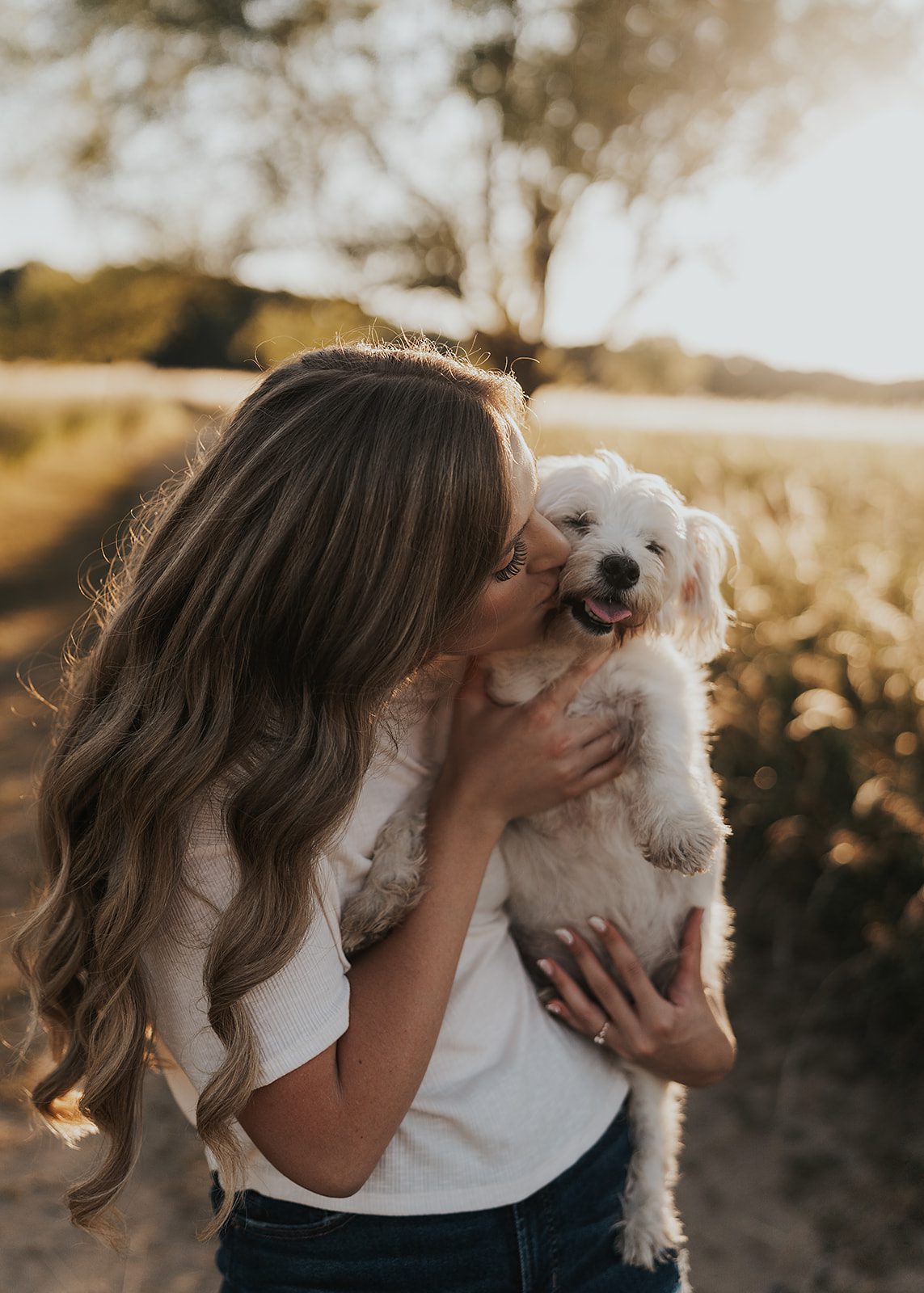 girl kissing her small white dog in a field during golden hour