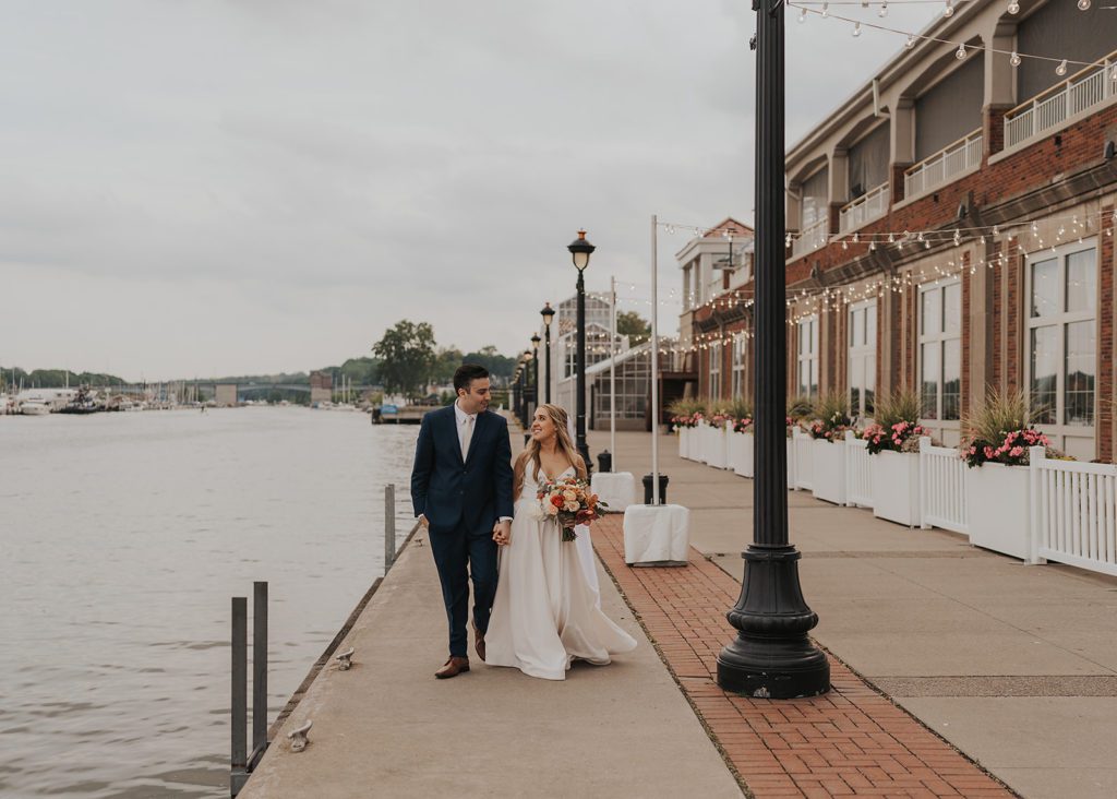 bride and groom waling down beside the river outside of the arbor at the port wedding venue in rochester ny