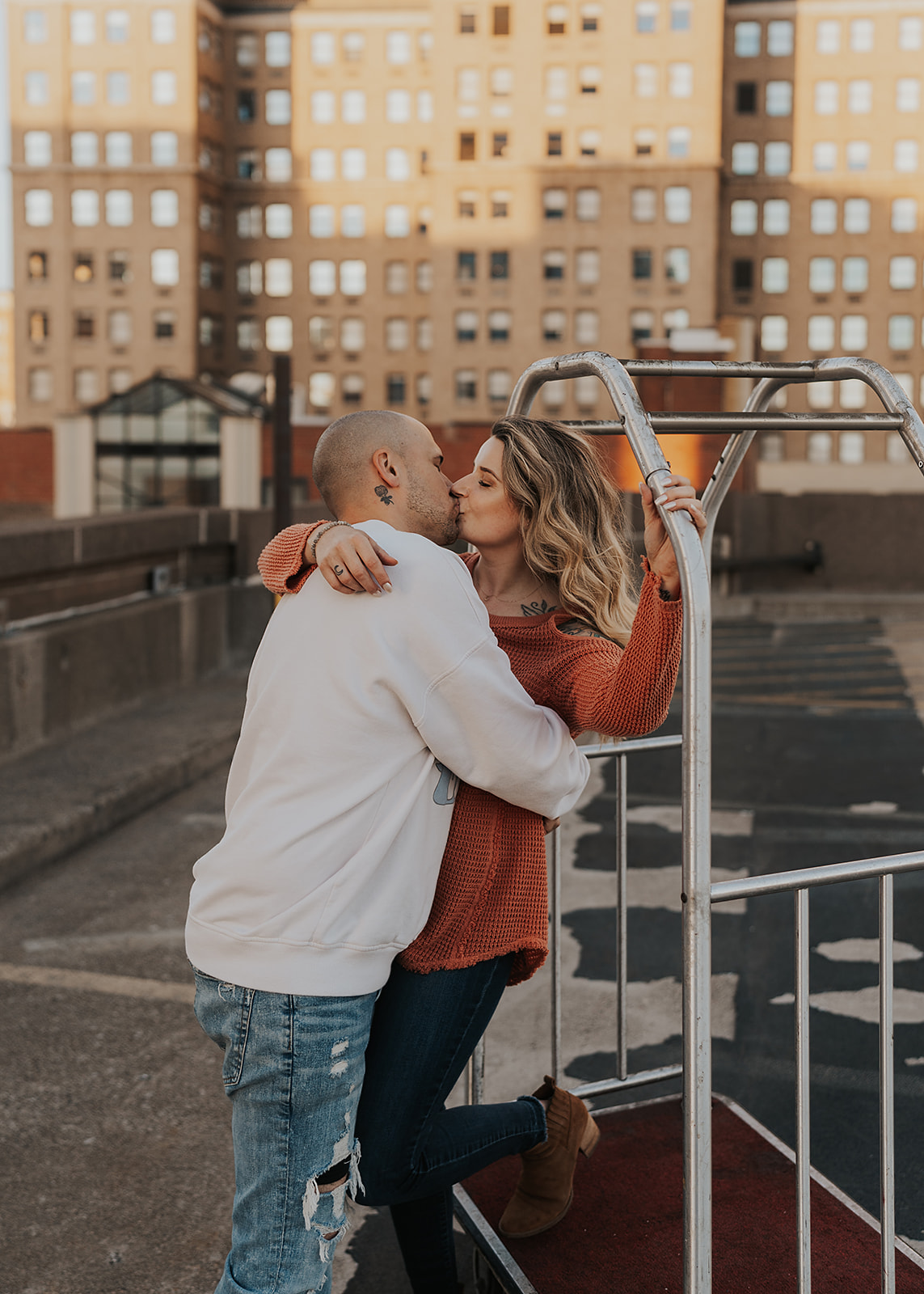 fiance kissing his girlfriends neck while standing on a bell hop cart on top of a parking garage