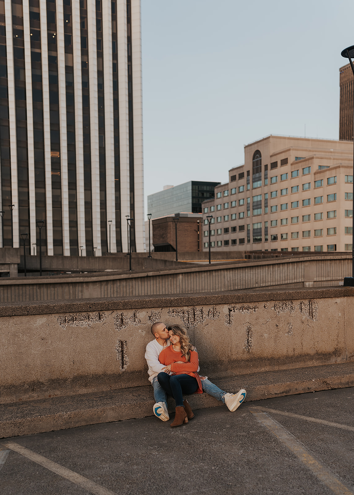 couple sitting on a parking garage roof top with the cityscape behind them for the engagement session