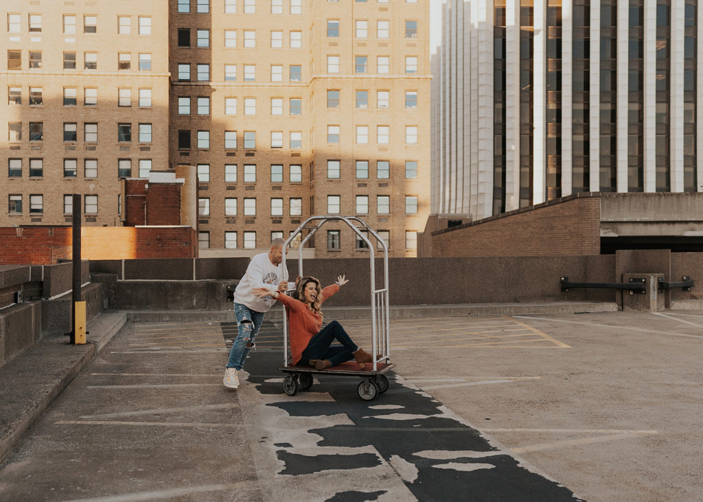 Fiance pushing his girlfriend in bell-hop cart on top of a parking garage during their engagement session