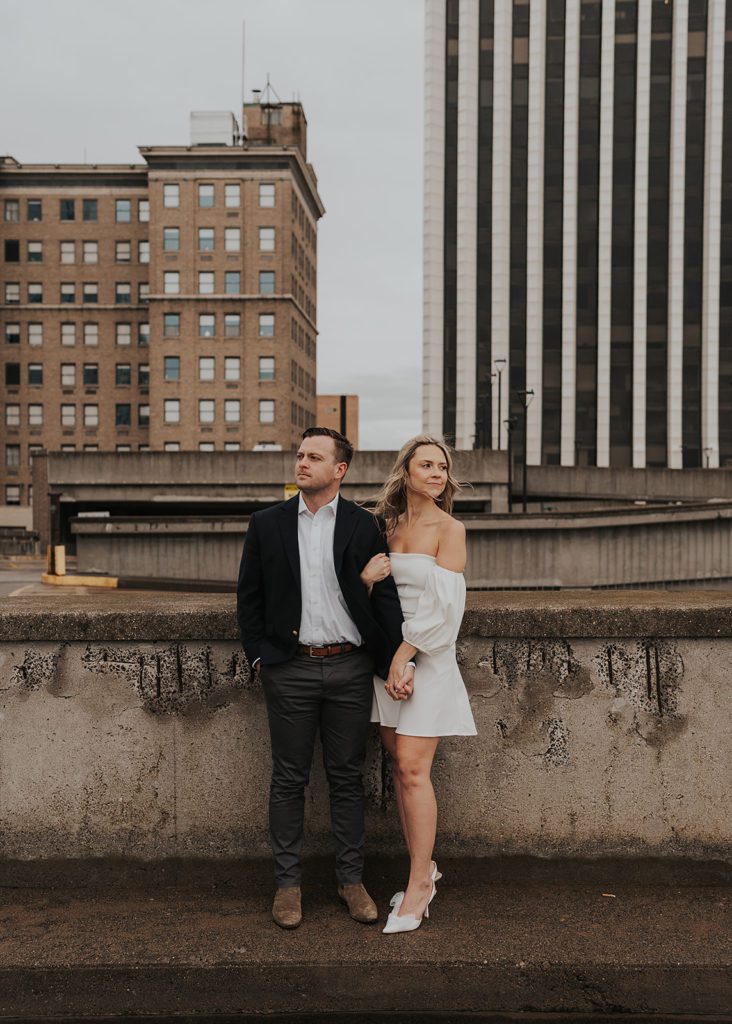 engaged couple holding hands on a parking garage rooftop in Rochester ny looking opposite directions with the cityscape in the background 