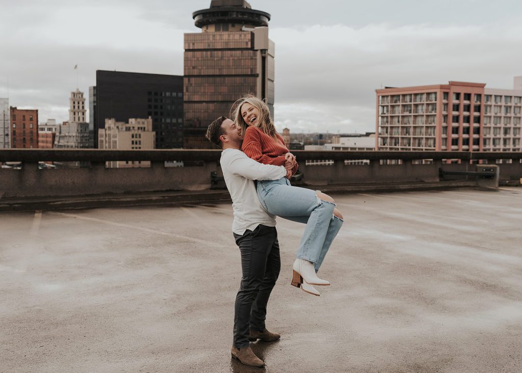 Fiance spinning his girlfriend around while she laughs during their engagement photos on a rooftop parking garage in Rochester NY