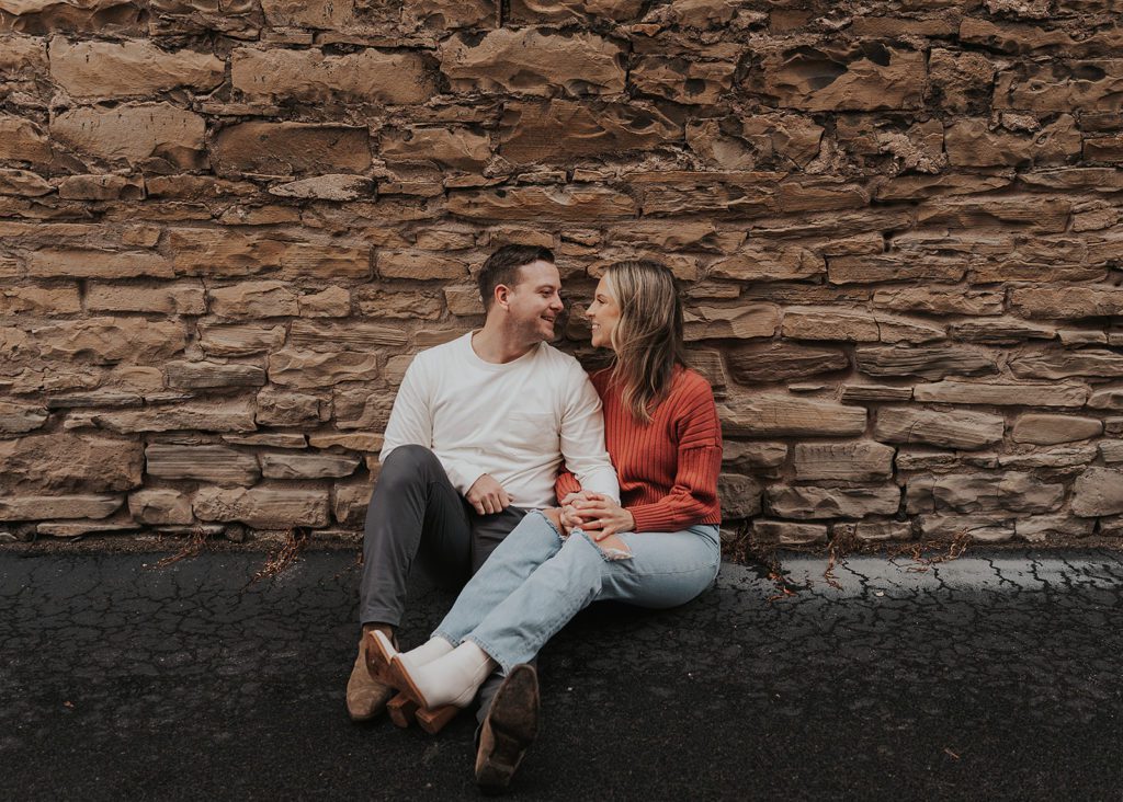 engaged couple sitting against a wall in an alley in Rochester NY while smiling at each other