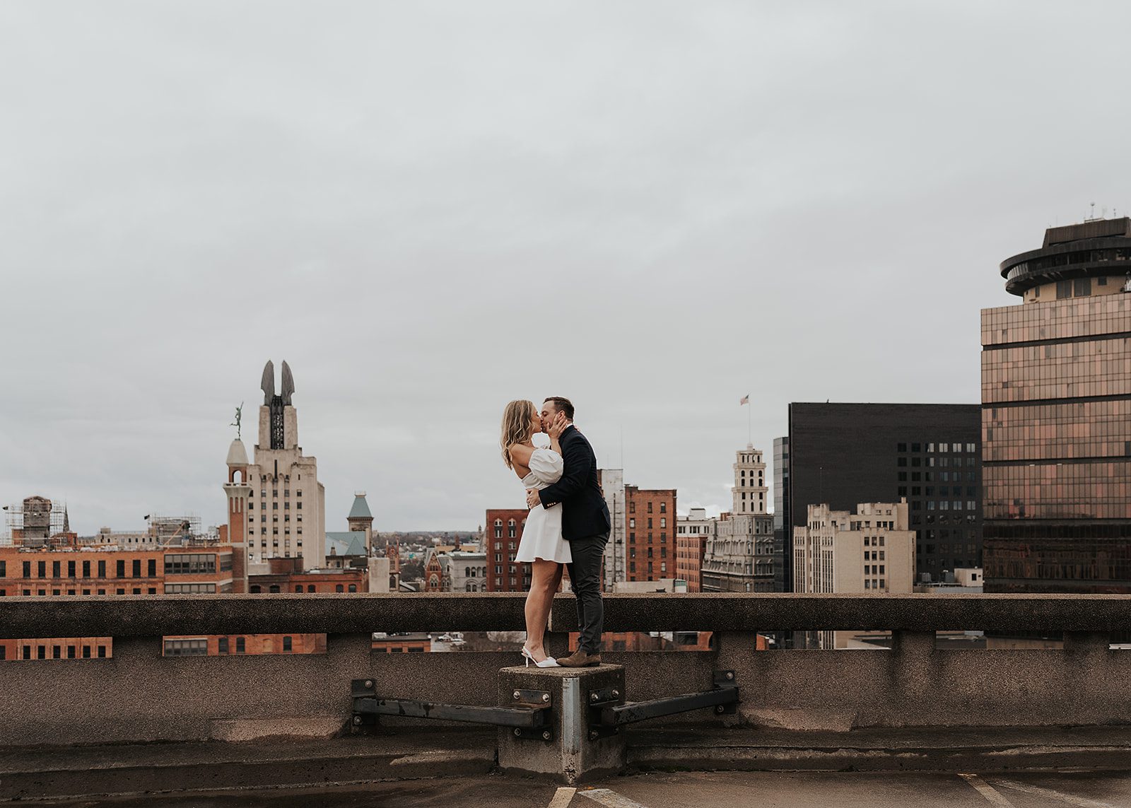 Engaged couple on rooftop in rochester NY kissing with the cityscape in the background