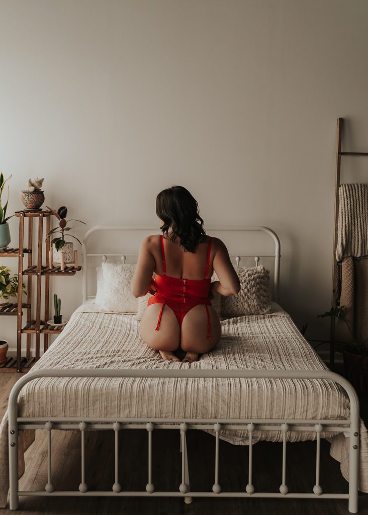 women sitting on the bed in red lingerie facing away from the camera during her boudoir session in Rochester NY at Gallery 48 photo studio