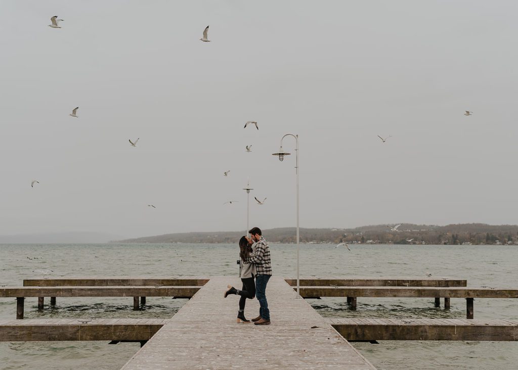 engaged couple standing on a pier in Rochester NY kissing while seagulls fly in the background