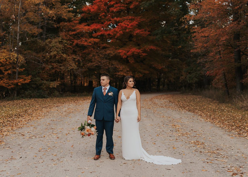 Bride and Groom holding hands while standing on dirt road in front gorgeous fall trees at Cummings Nature Center a Rochester NY Wedding Venue