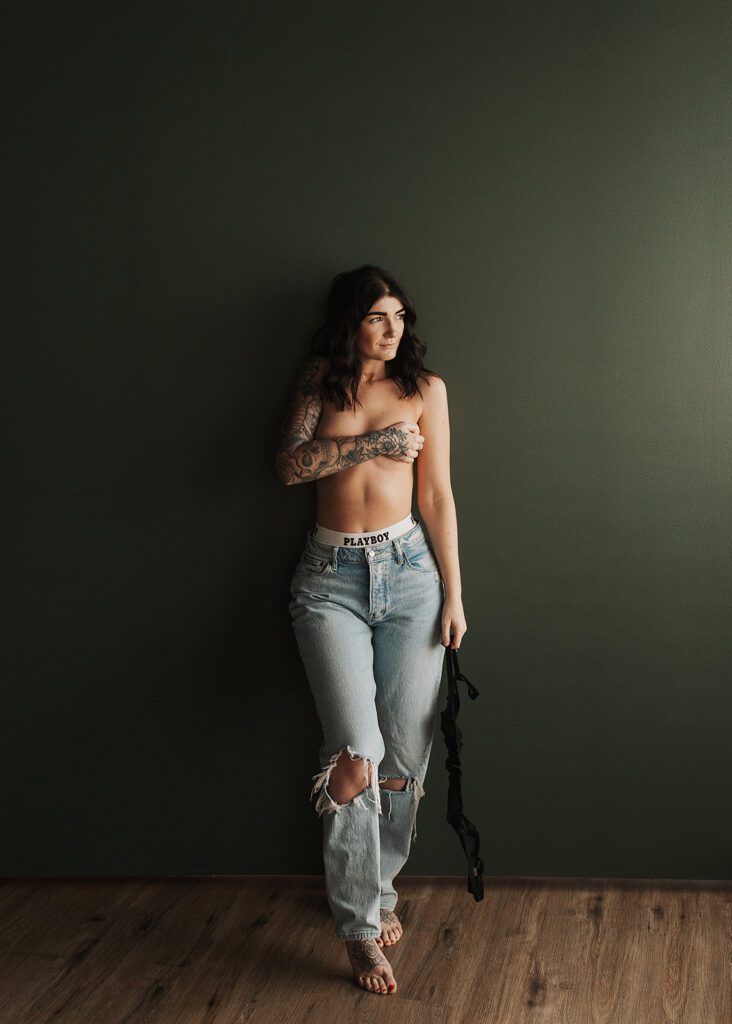 brunette women wearing jeans and covering her breasts leaning up against a wall during her rochester ny boudoir photoshoot at gallery 48 photography studio