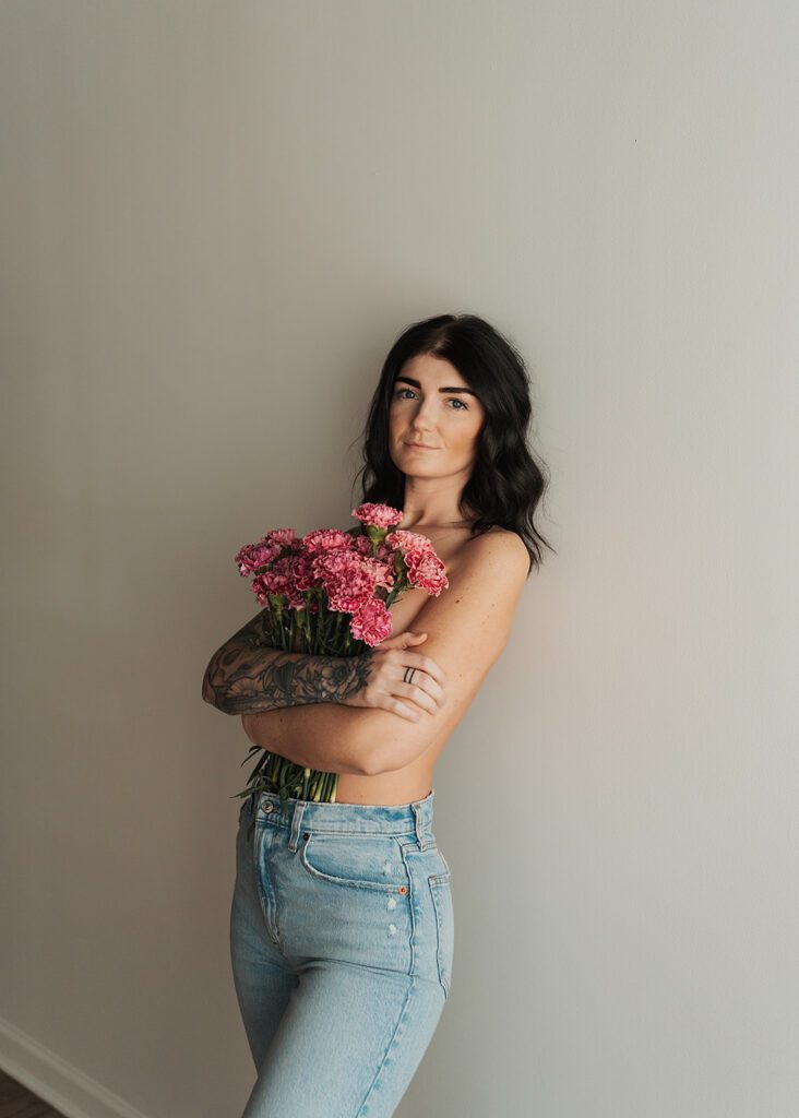 tattooed brunette women wearing jeans and holding flowers over her breasts while she leans against a wall in gallery 48 photography studio during her rochester ny boudoir photoshoot