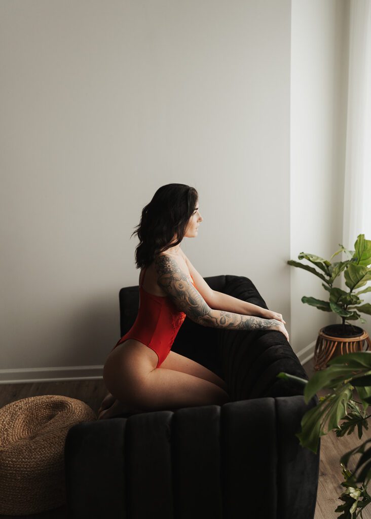 tattooed brunette girl wearing red lingerie sitting on her knees on a green velvet couch looking out the window during her rochester ny boudoir photoshoot at gallery 48 photography studio