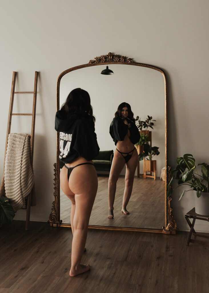 brunette women standing in front of a mirror looking at herself while wearing a sweatshirt and thong during her rochester ny boudoir photoshoot at gallery 48 photography studio