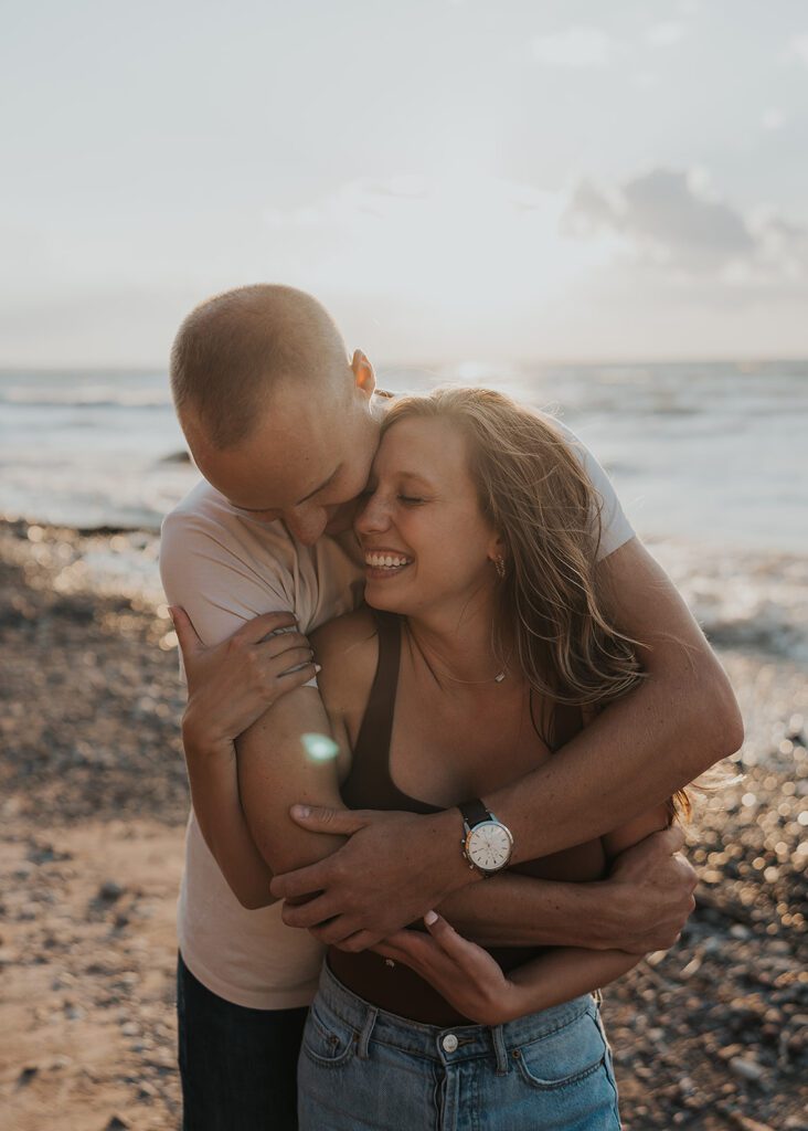 fiance hugging his girlfriend from behind on the beach while he nuzzles into her neck and she smiles and laughs