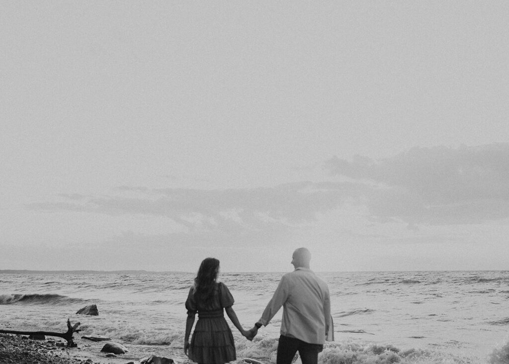 engaged couple holding hands and walking down the beach away from the camera during their beach engagement photoshoot