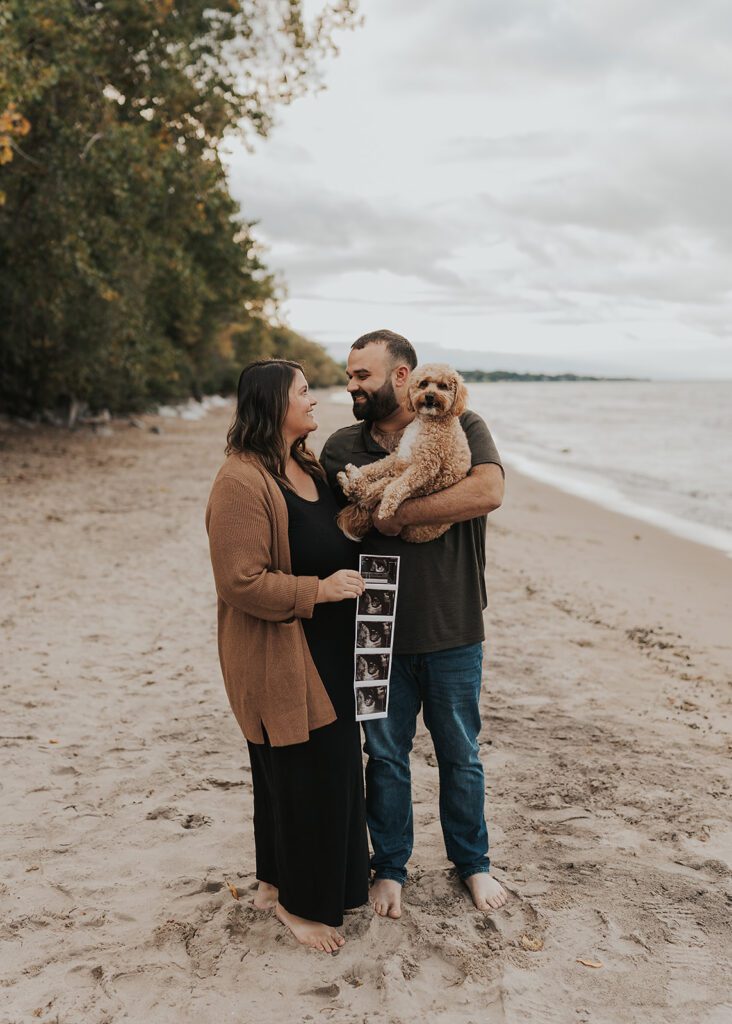 married couple with husband holding their dog and wife holding a baby sonogram on the beach in rochester ny during their pregnancy announcement photoshoot