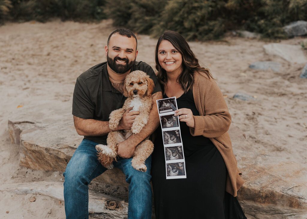 married couple with their dog sitting on the beach with the wife holding a baby sonogram during their pregnancy announcement photoshoot