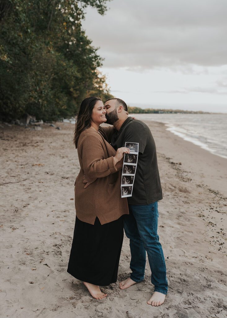 husband kissing wife on the cheek while she holds a baby sonogram up to the camera on a beach in rochester ny during their pregnancy announcement photoshoot