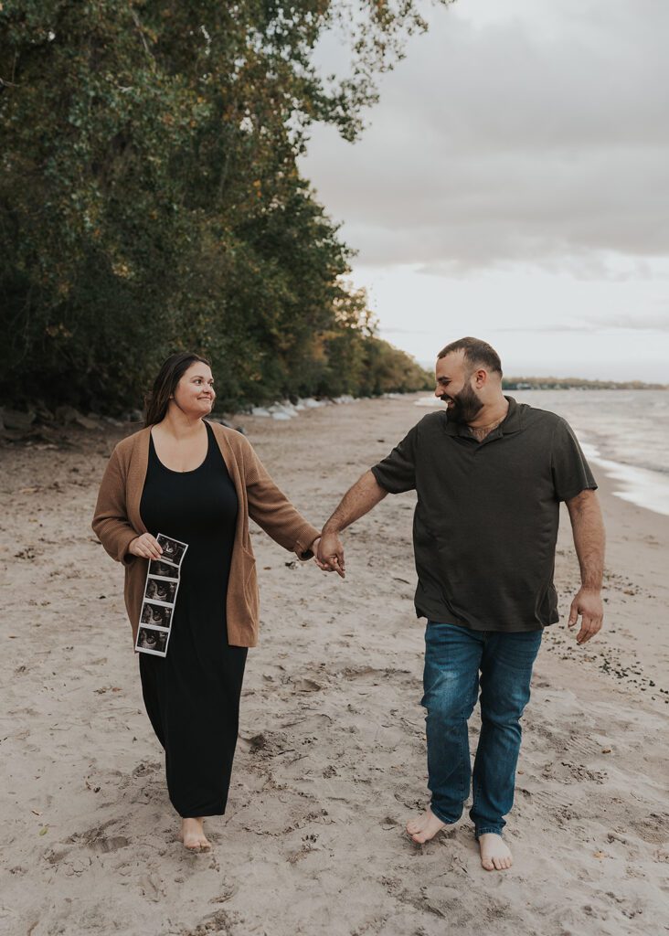 couple walking on the beach holding hands with wife holding a baby sonogram in her hands during their rocchester ny pregnancy announcement photoshoot