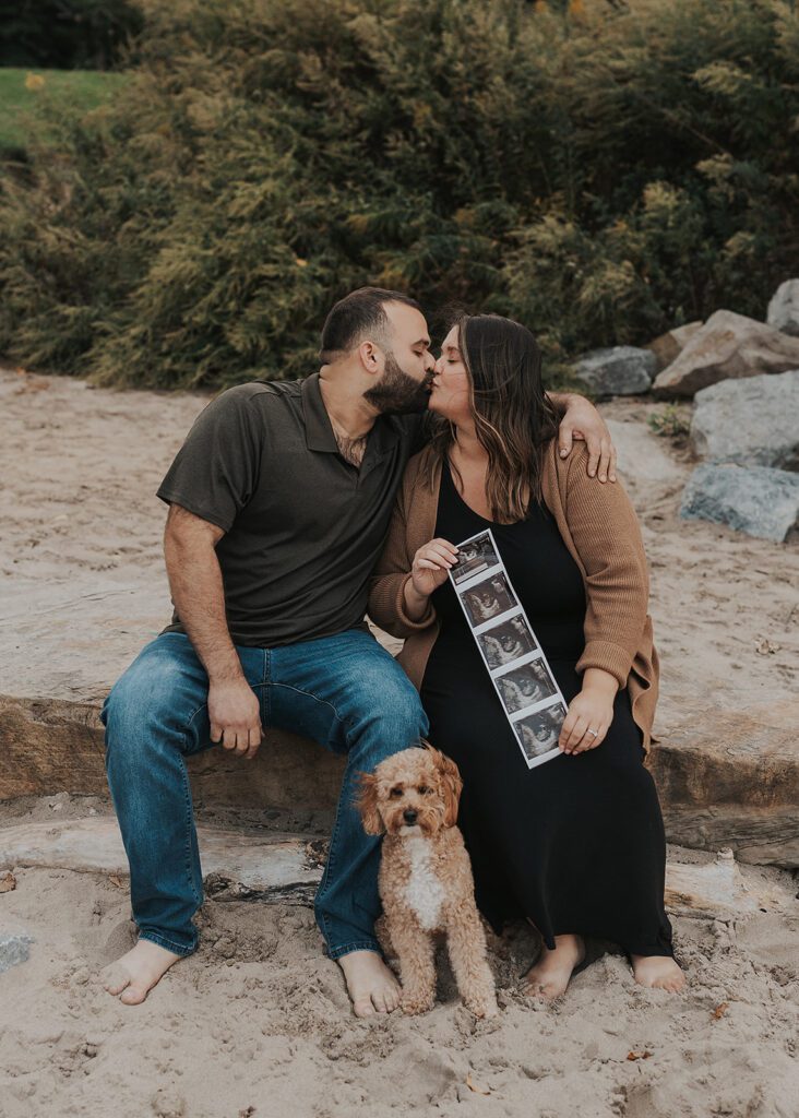 couple kissing while holding up their baby sonogram on the beach in rochester ny during their pregnancy  announcement photoshoot 
