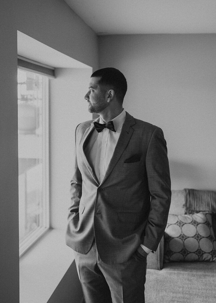 black and white image of groom standing in the window light while getting ready for the wedding ceremony