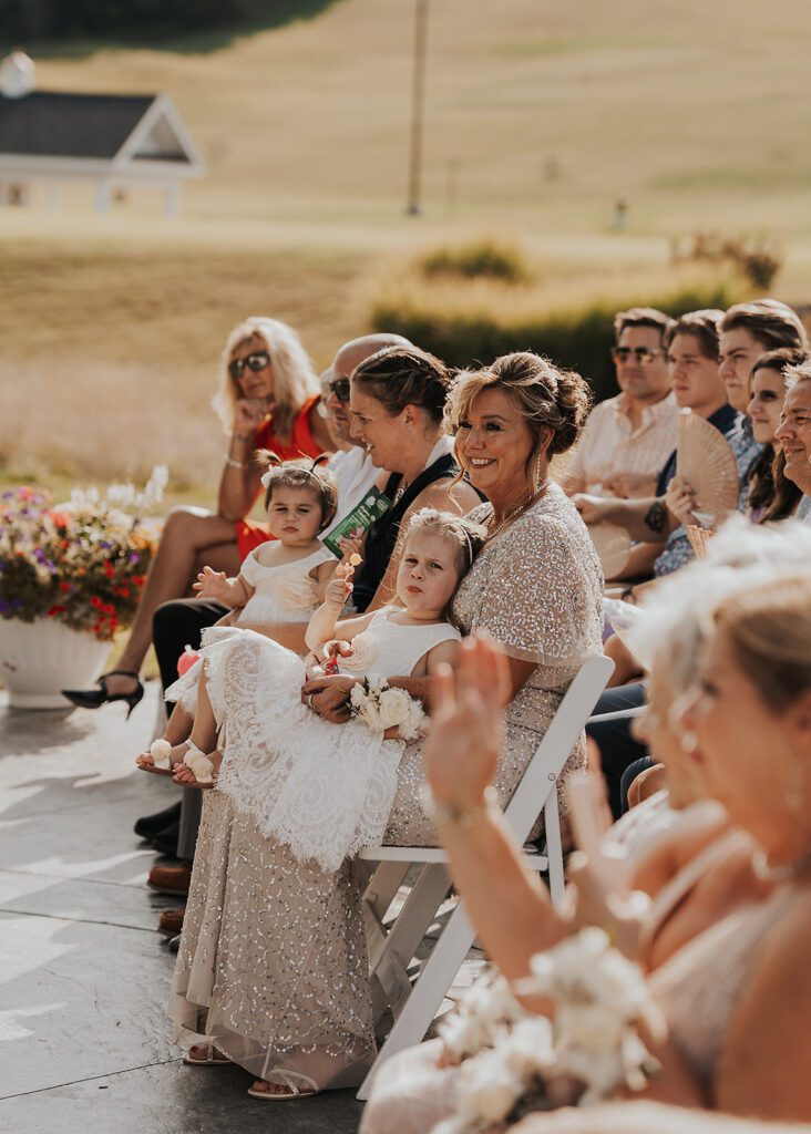 mom holding a little kid on her lap while she smiles at bride and groom during their wedding ceremony