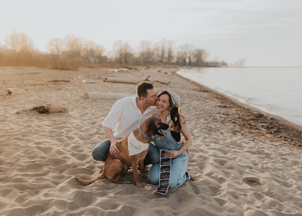 boho couple sitting on the beach with their dog while holding a baby sonogram photo announcing their pregnancy 