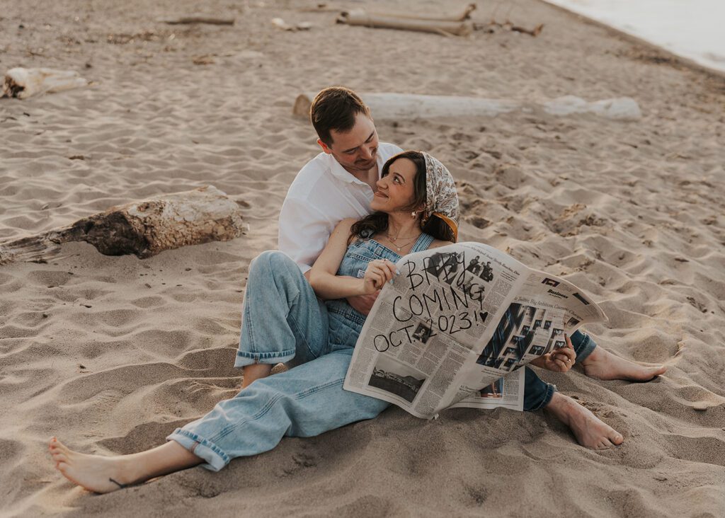 boho couple sitting on the each smiling at each other holding a newspaper announcing their baby during their pregnancy announcement photoshoot