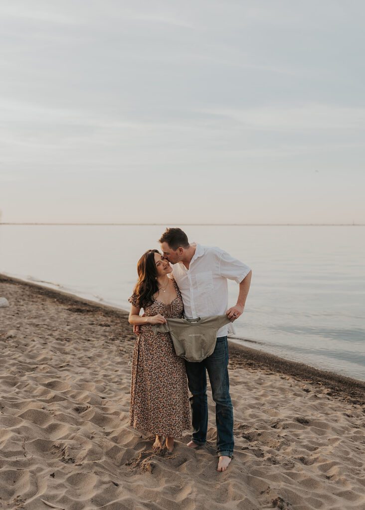 cute couple holding baby clothes on the beach during their pregnancy announcement in colombus ohio