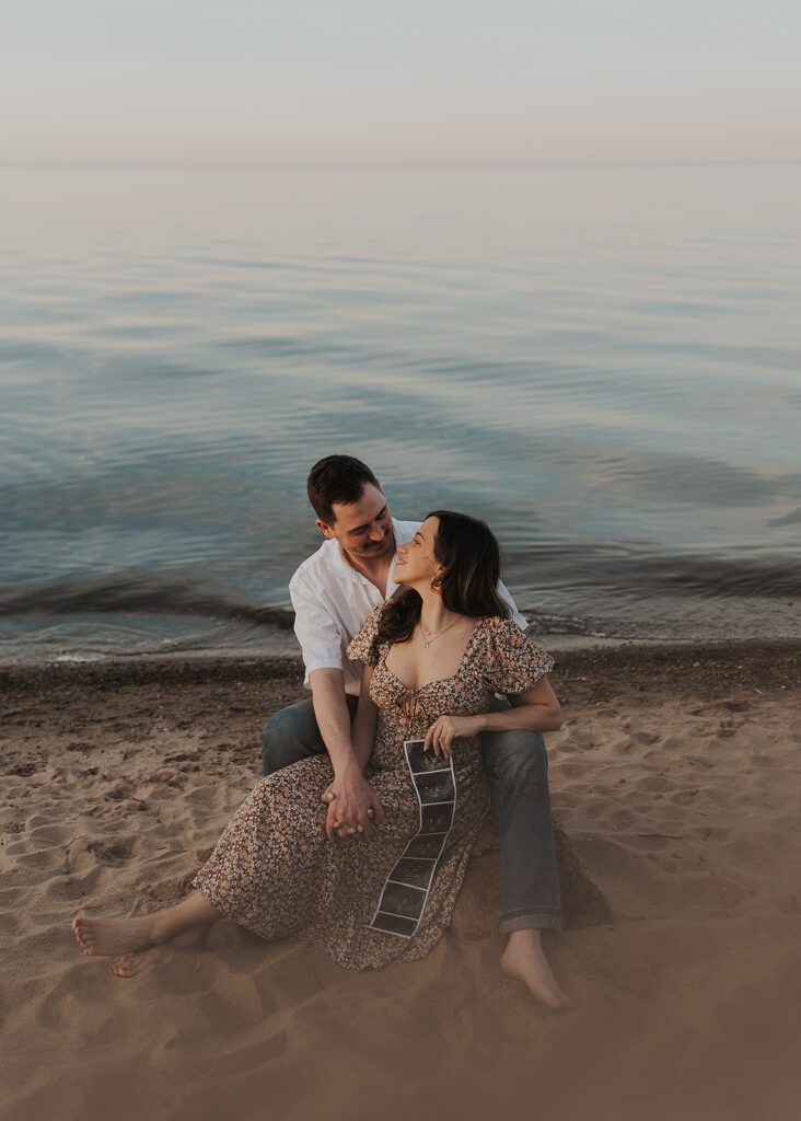 beachy boho couple sitting on the beach holding a baby sonogram during their pregnancy announcement photoshoot in colombus ohio
